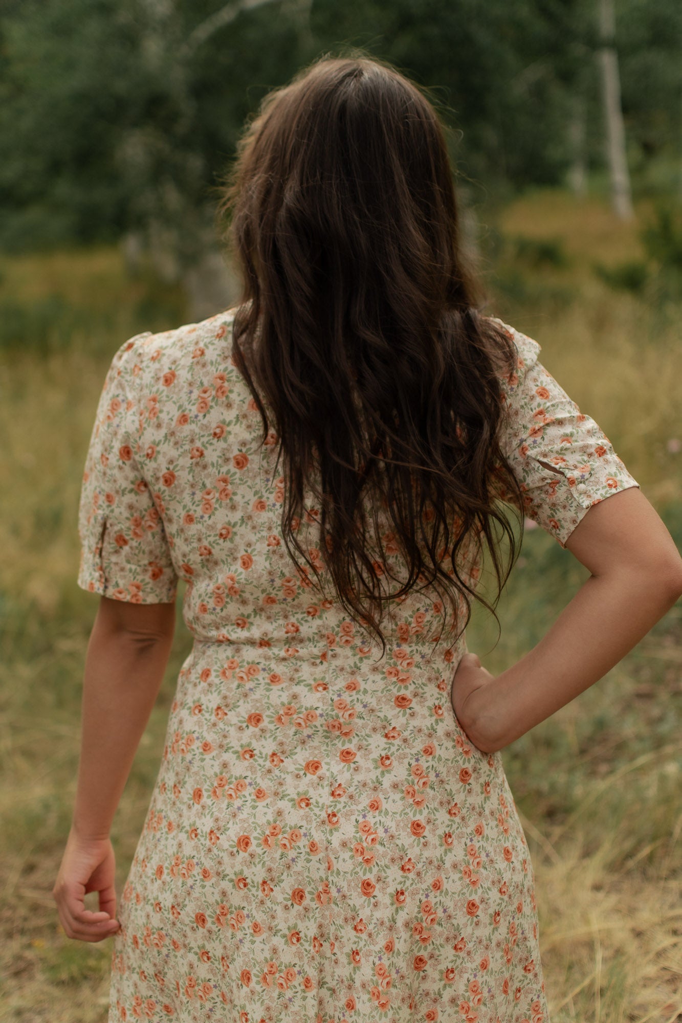 Woman in the woods wearing a floral dress, cream with peach roses, turned around revealing back dress details.