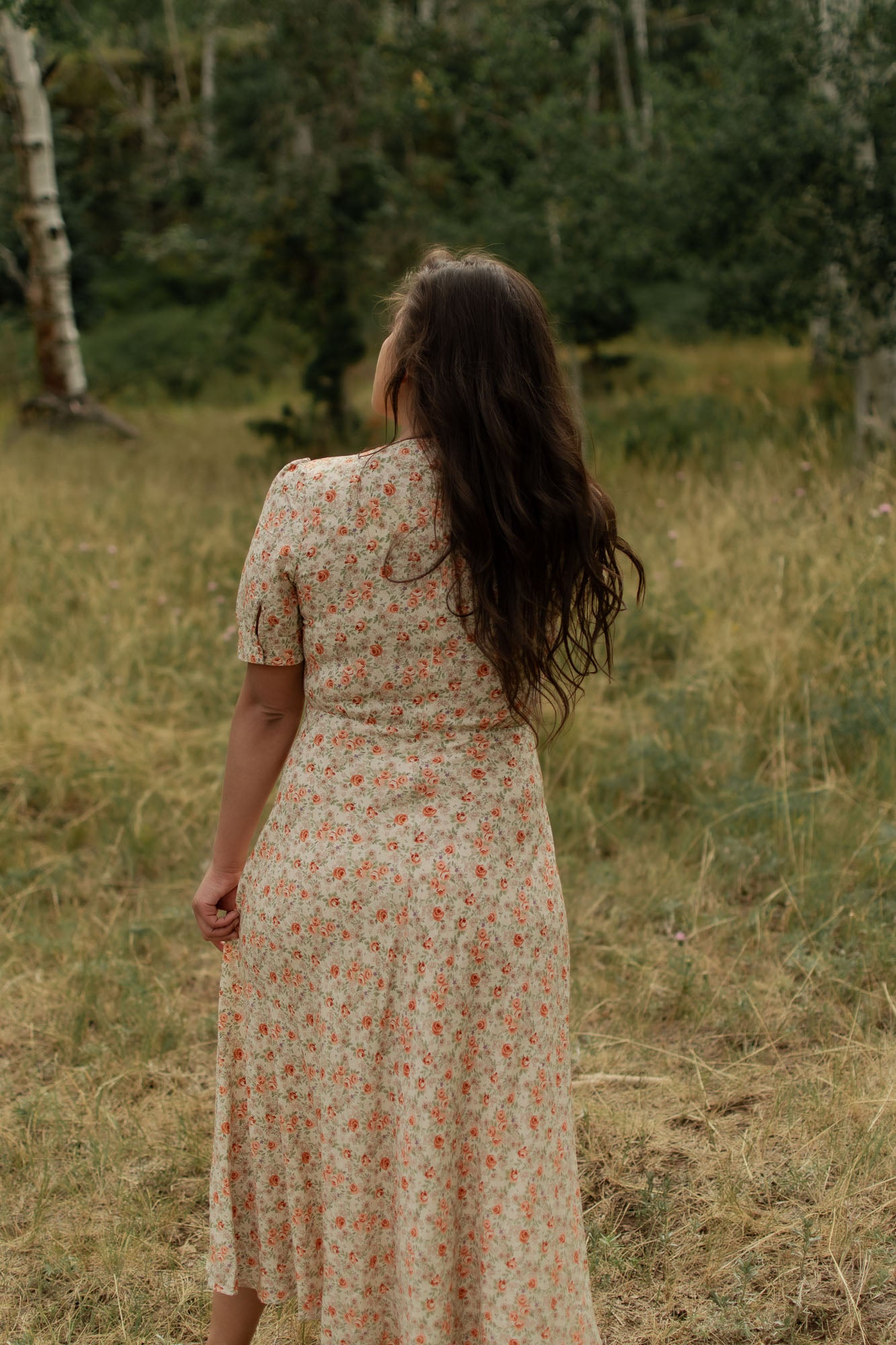 Woman in the woods wearing a floral dress, cream with peach roses, turned around revealing back of dress.