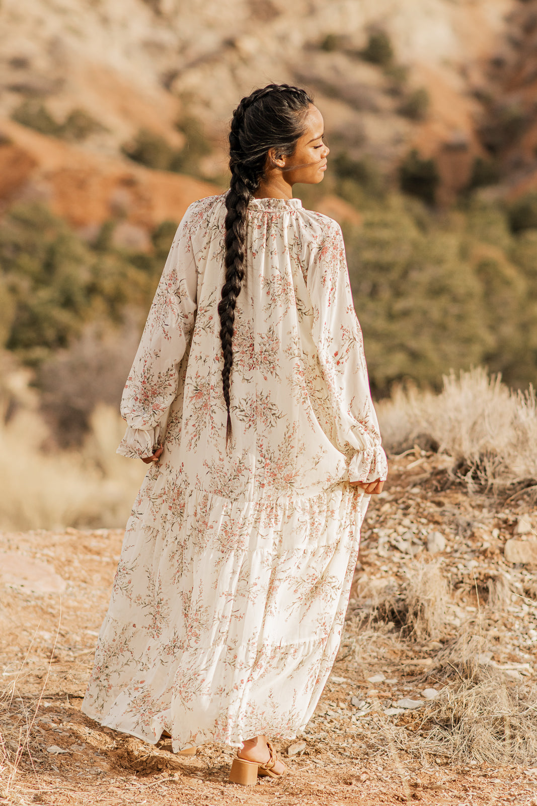 Women facing backwards displaying a elegant flowy, floral, cream, boho style maxi dress, lined, with bell cuff sleeves, and tiered bottom. bella by grace co.