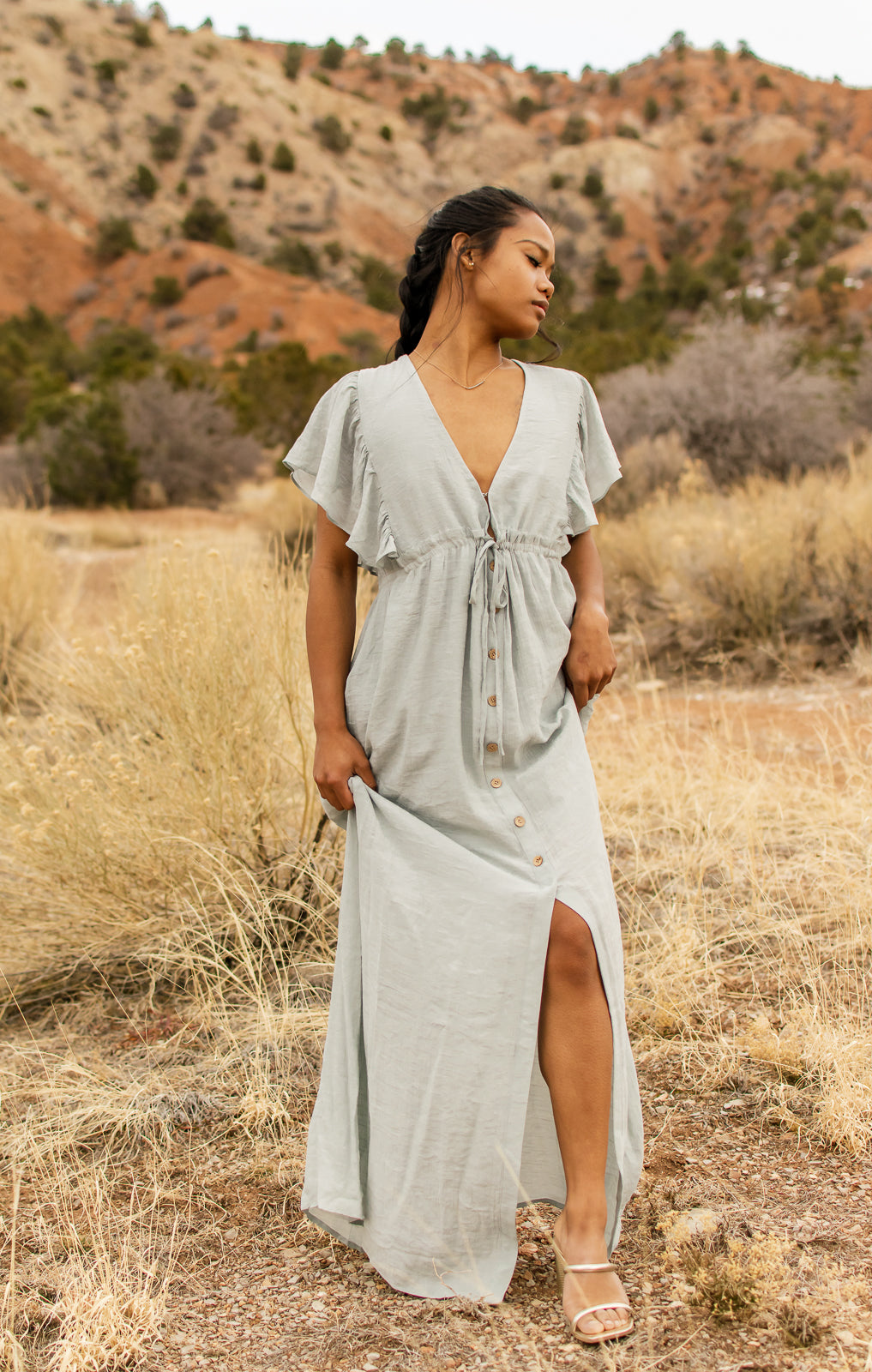 Women is modeling a light blue, V-neck, button down following a slit, with angel sleeves, maxi dress. bella by grace co.